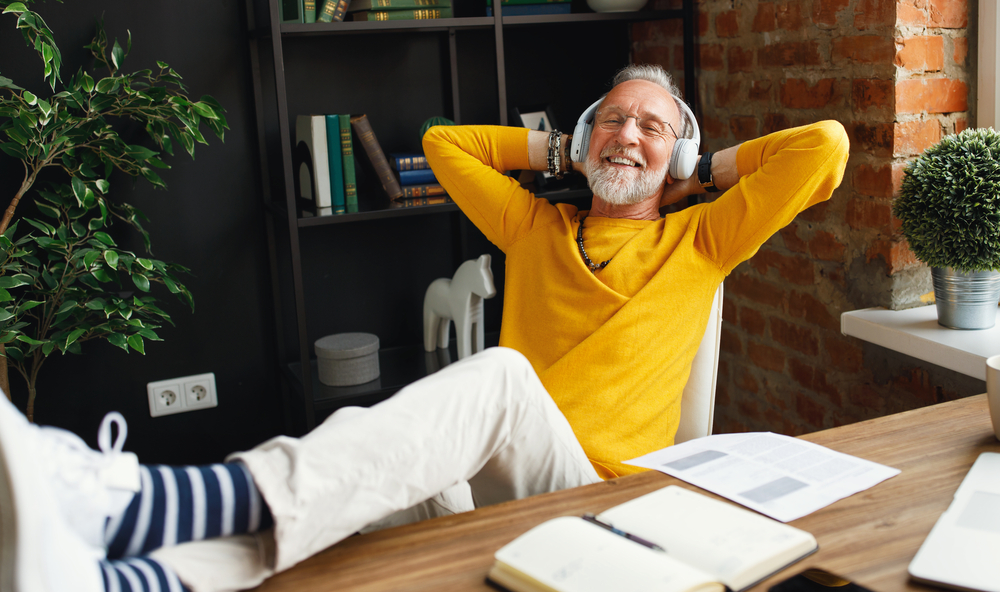 A senior man puts his feet up on the desk and listens to his over the ear headphones